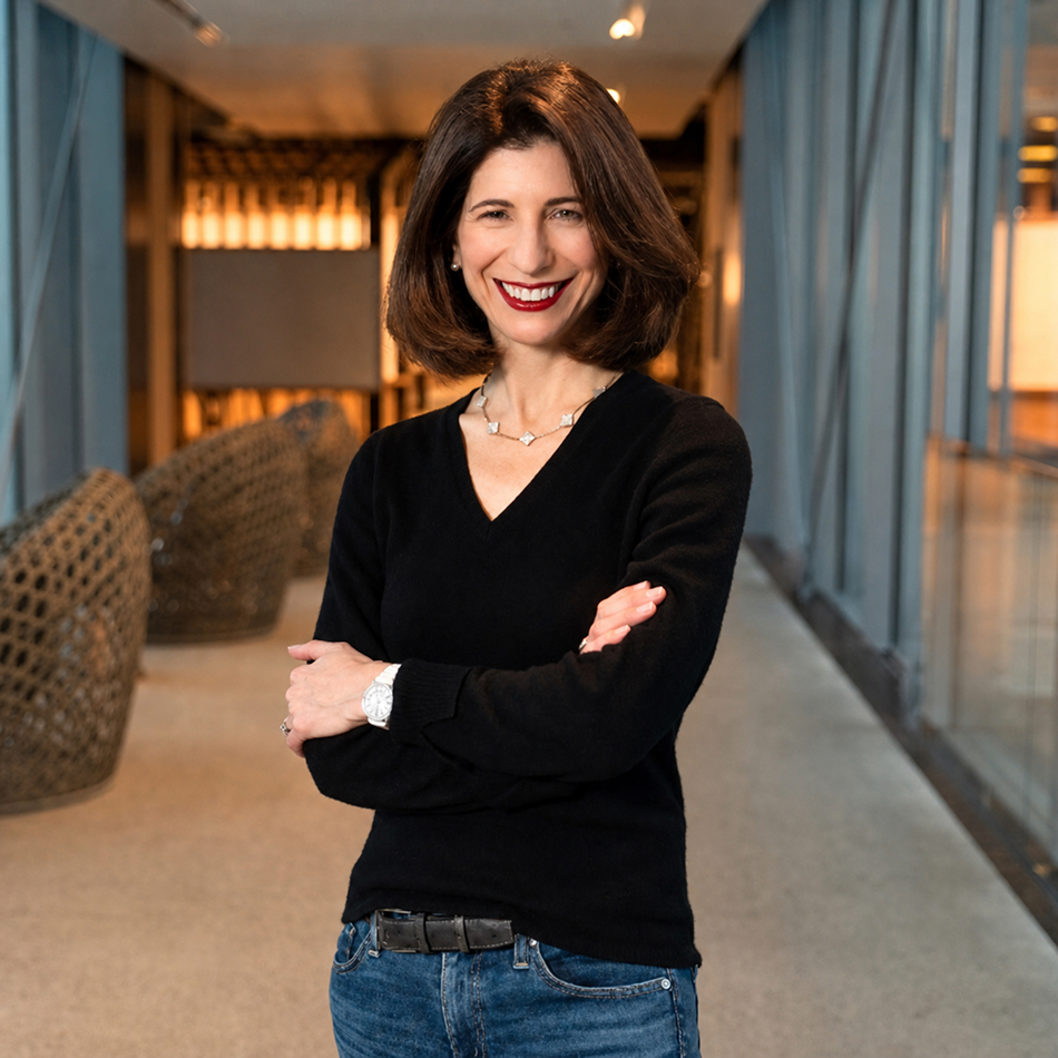 Ariane Gorin smiling with arms crossed in a modern, glass-walled office corridor.