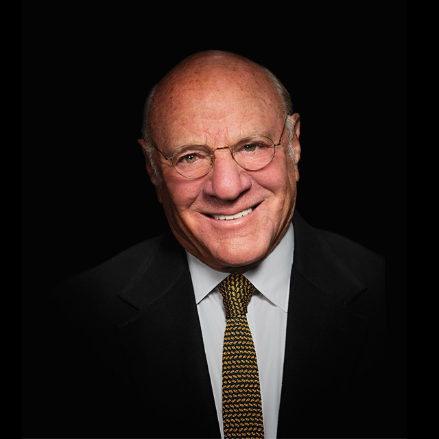 Barry Diller smiling and wearing glasses, in a dark suit, a white shirt, and a patterned tie, set against a black background.