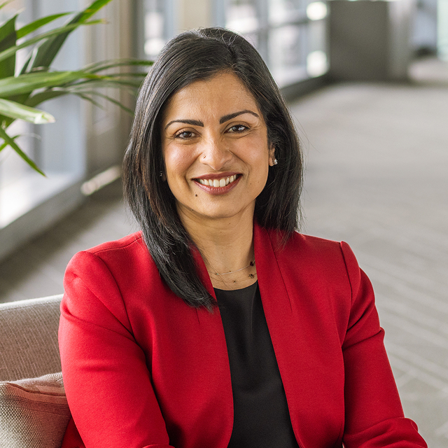 Shilpa Ranganathan smiling and wearing a red blazer over a black top, seated on a light chair in a bright hallway with a plant in the background.