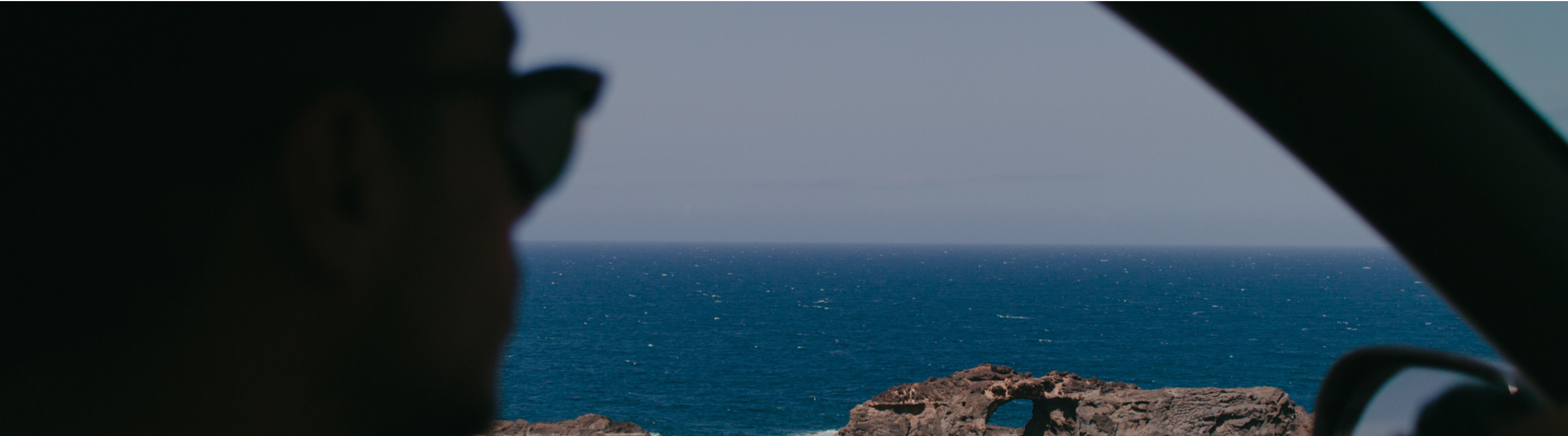 A side-profile silhouette of a person wearing sunglasses seated inside a vehicle, gazing out at a bright blue ocean and rocky coastline framed by the car’s window and A‑pillar.