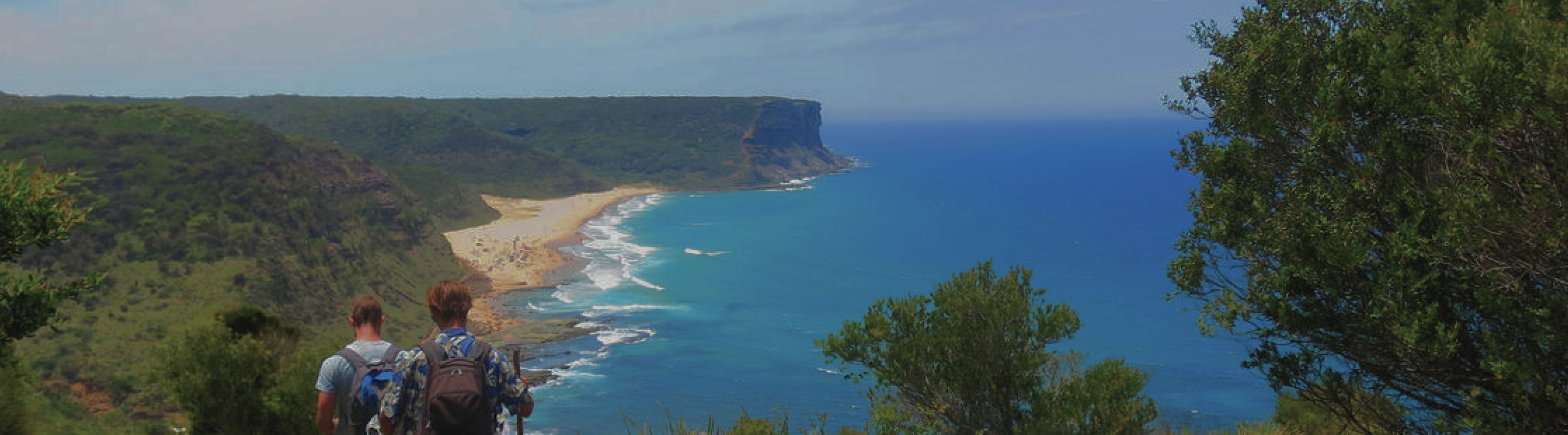 Two young men trekking through a national park with sticks and backpacks on with a view of the beach ahead on top of a hill.