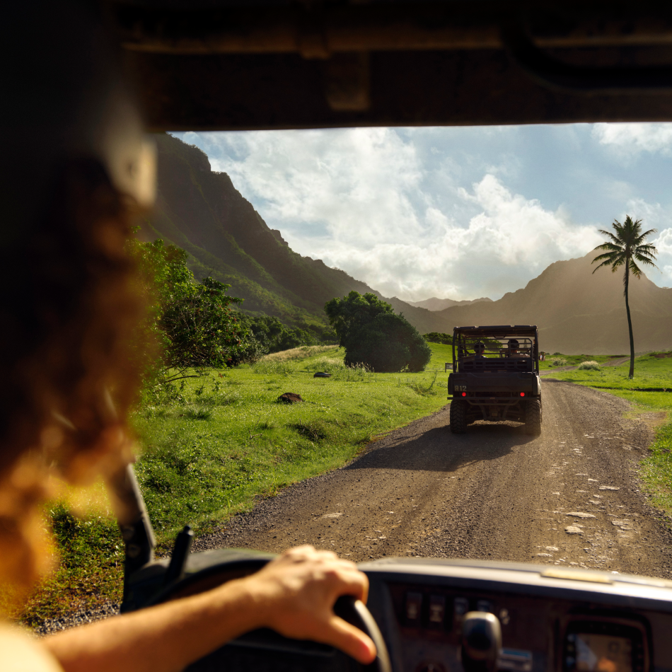 View through the windshield of a person driving on a road following another vehicle at Kualoa Ranch, featuring green grasslands, a palm tree, mountains in the distance and a cloudy sky above.