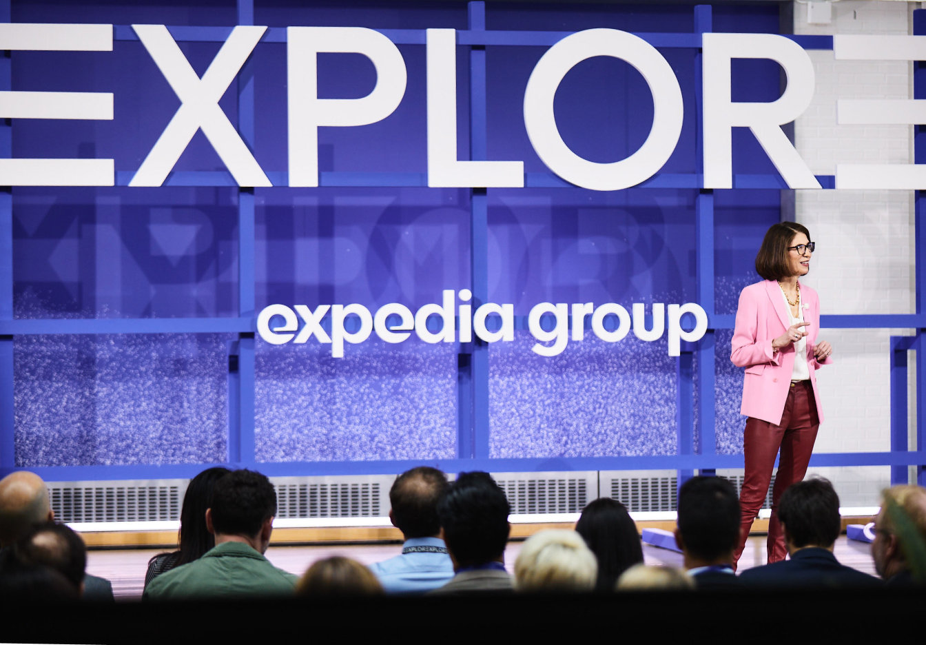 Speaker presenting on stage at Expedia Group event with audience in foreground and large “explore” sign in background