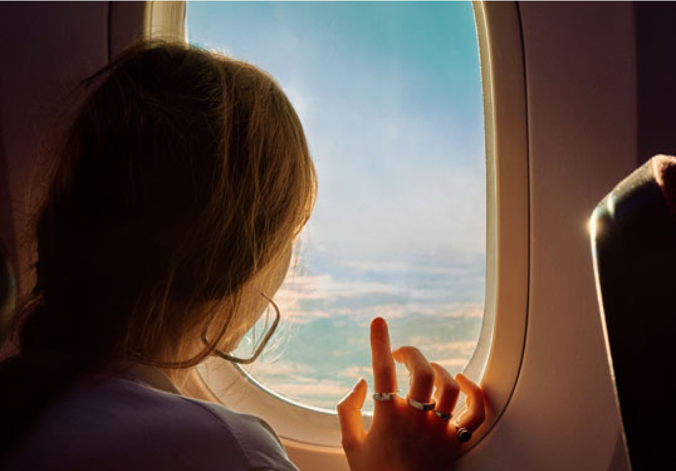 A young woman looks out an airplane window pointing to the clouds and sky from a dark exterior of the plane