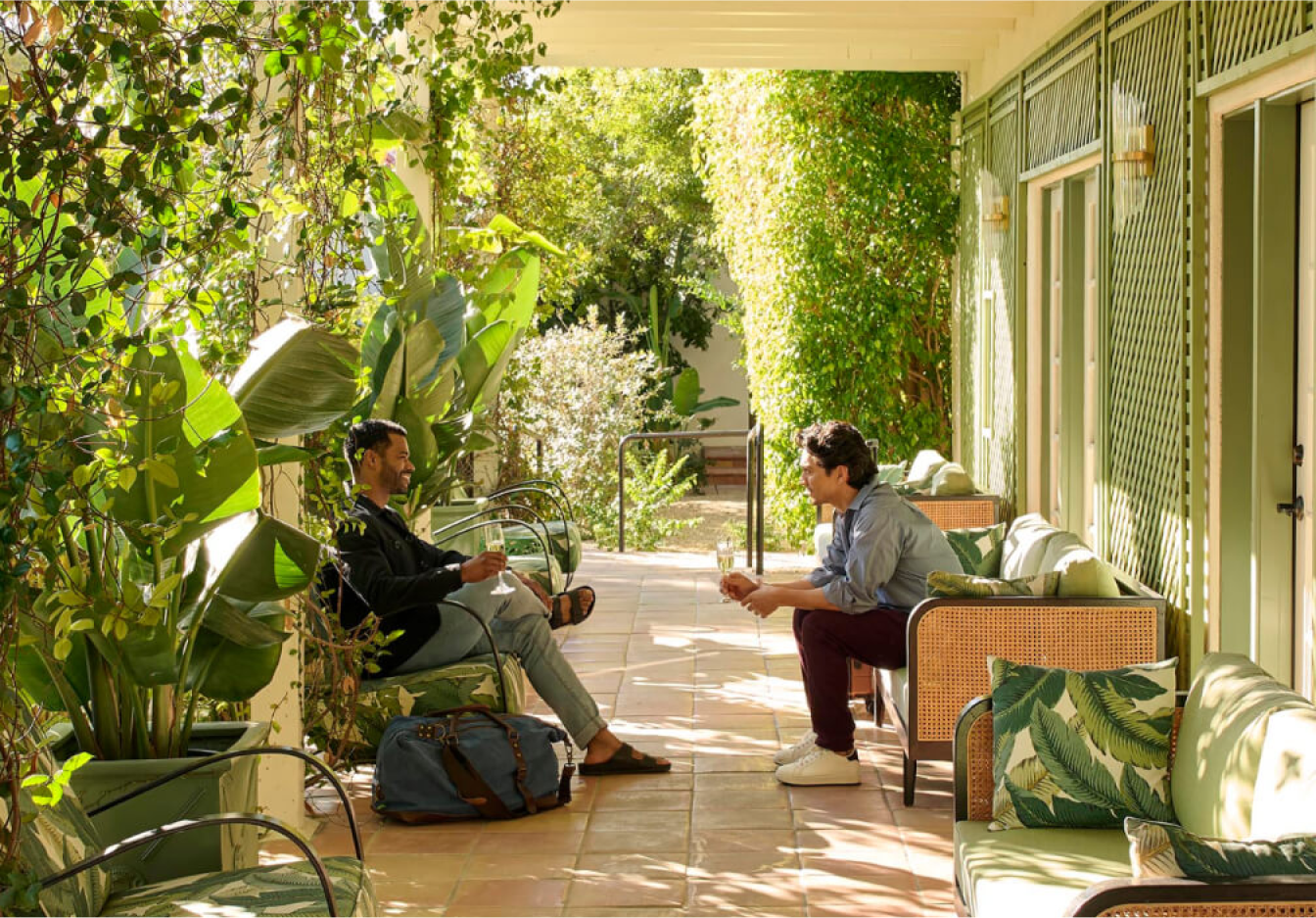 Photograph of two men seated in a hotel courtyard surrounded by lush greenery, engaged in conversation with their luggage nearby.