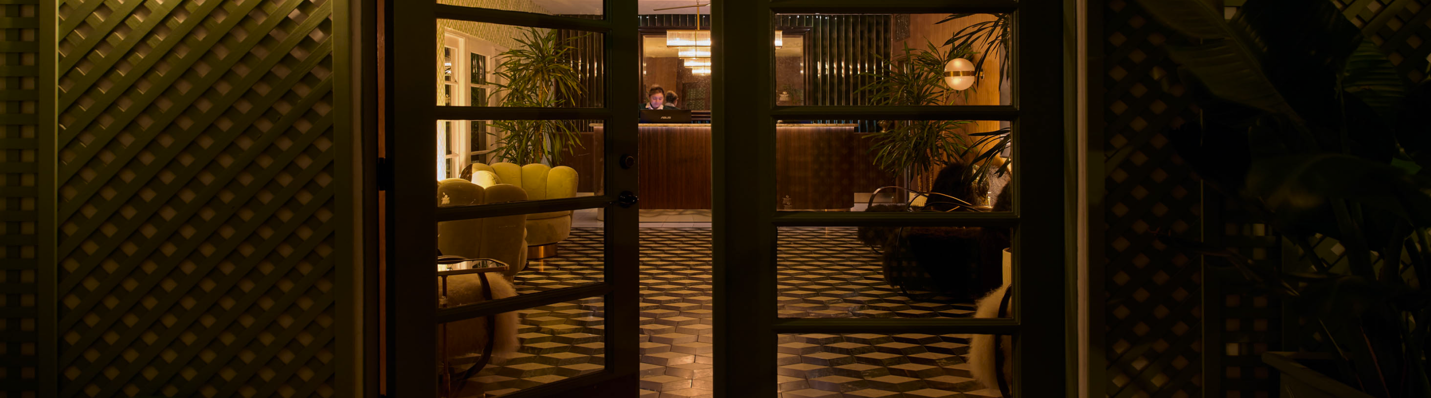 A view of the entrance to the hotel's lobby with the left door slightly open, revealing a hotel staff behind the front desk.