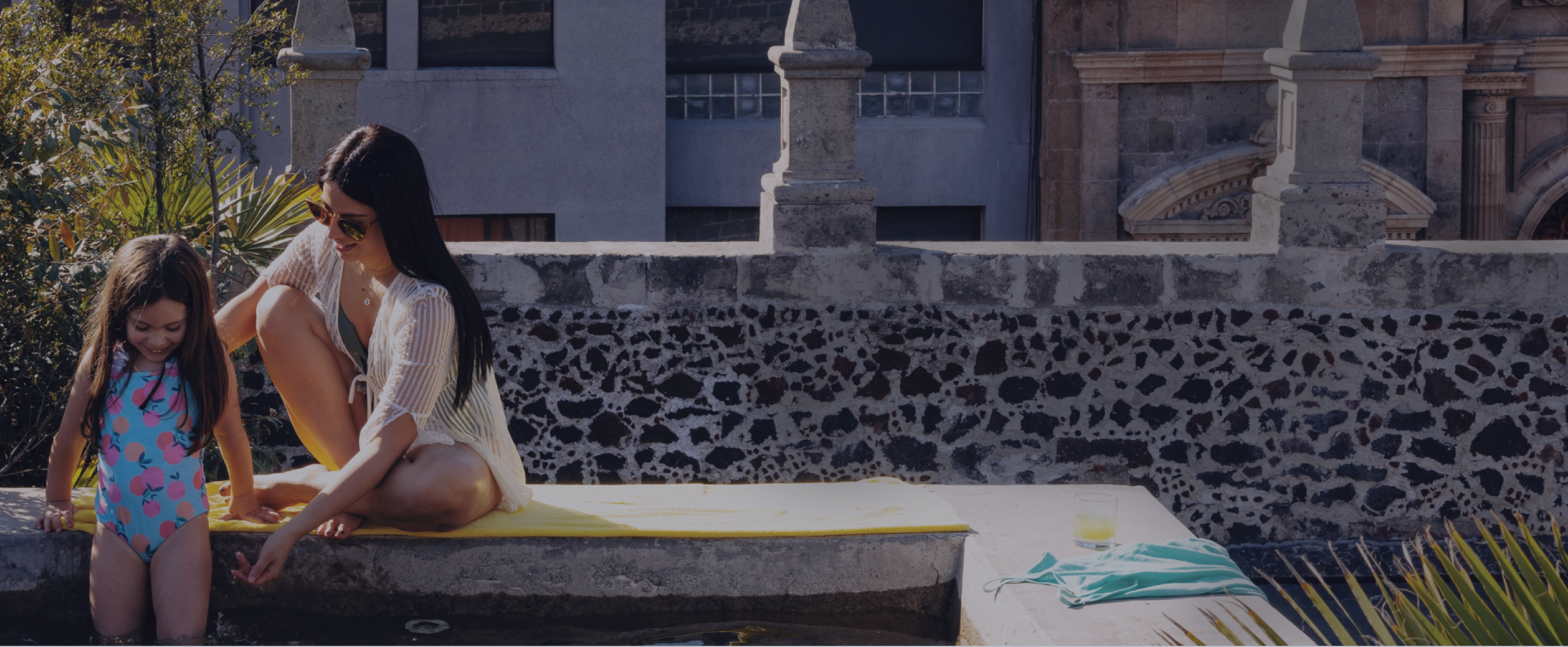  A mother and daughter sit at the edge of a courtyard pool, dipping their feet in the water together.