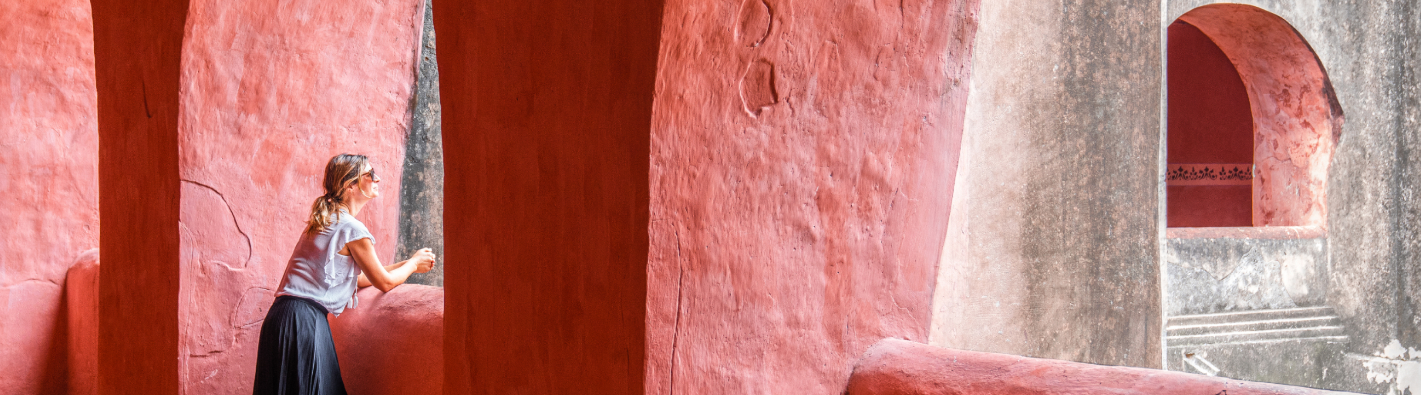 A woman leans on a red stone balcony, gazing thoughtfully through arched openings of an ancient, weathered structure with curved walls and soft natural light.