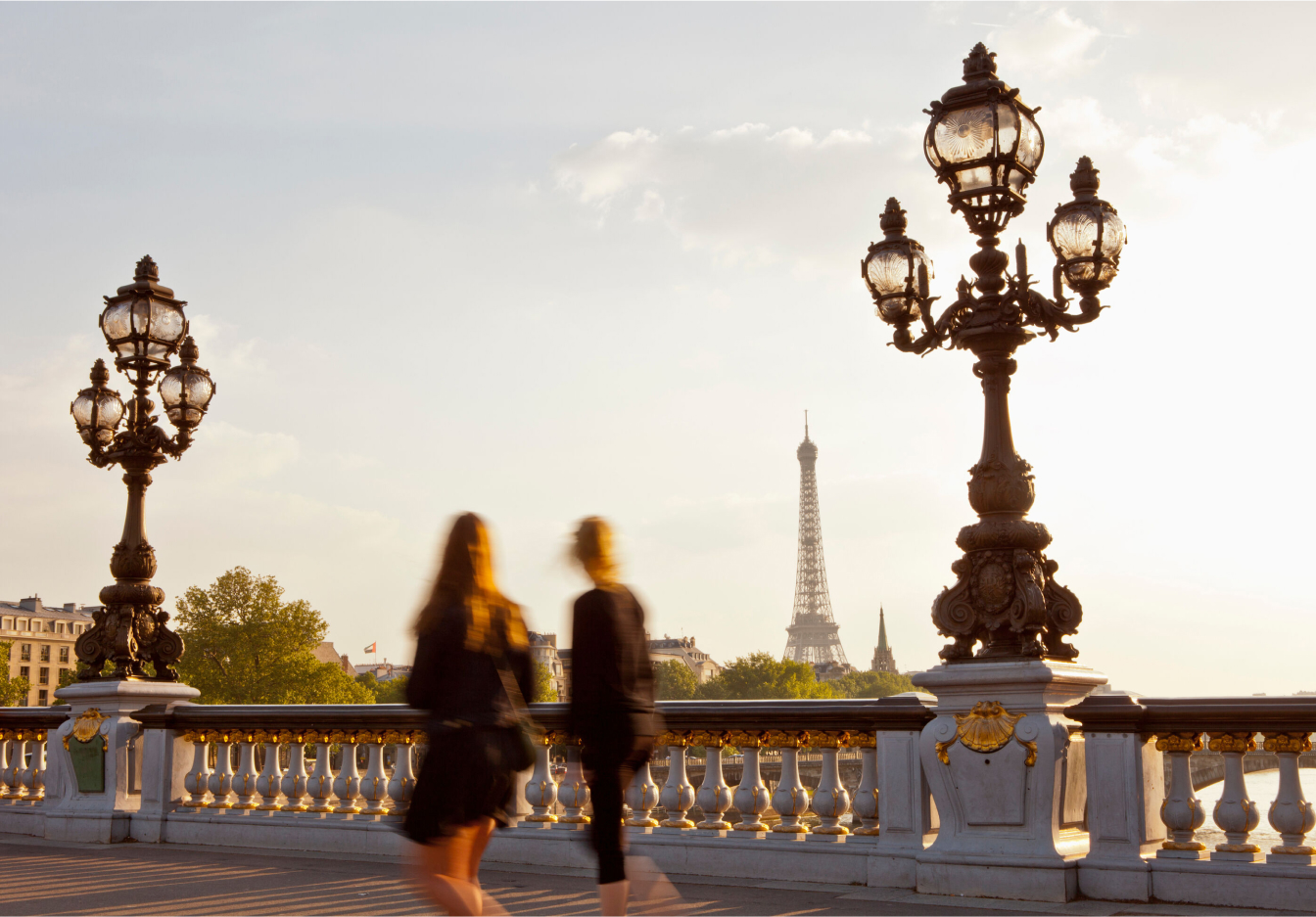 Two women walking along a historic bridge in Paris, with the Eiffel Tower in the distance.