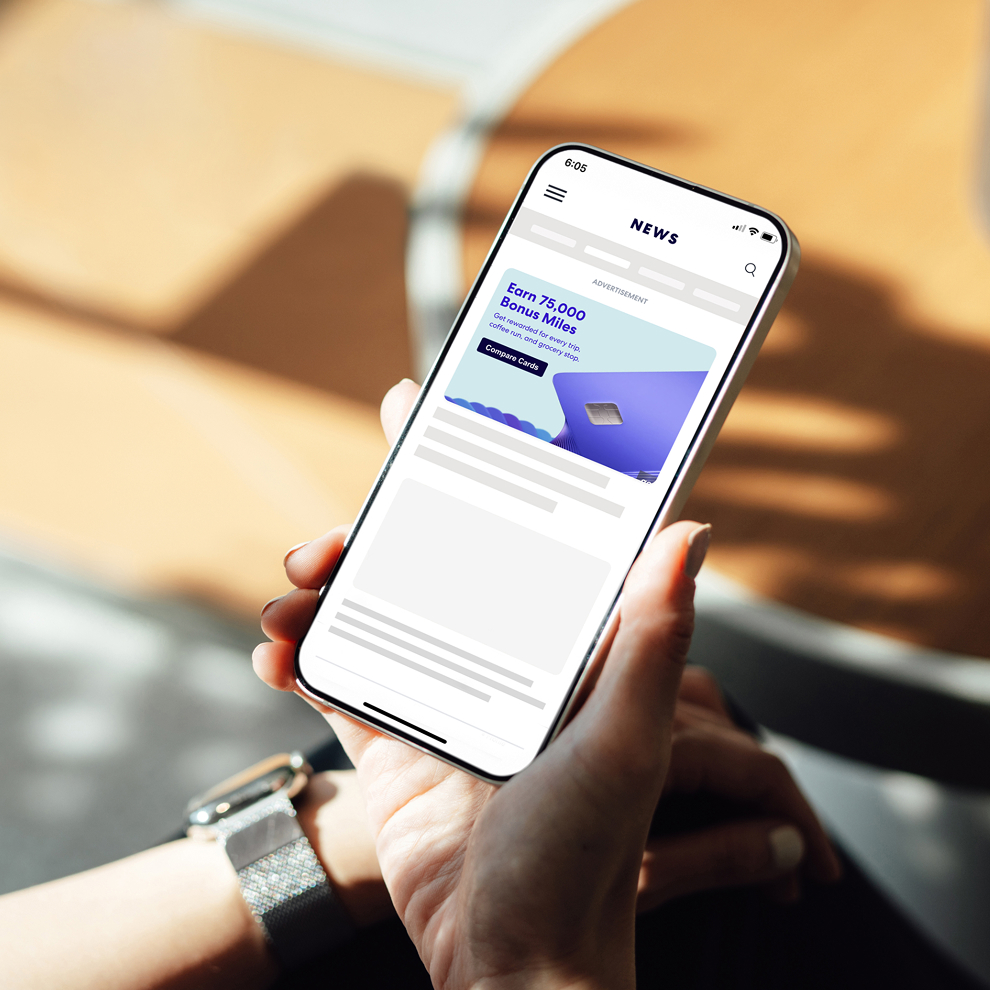 Close-up of a hand holding a smartphone displaying a newsfeed-style mobile page with a sponsored “Earn 75,000 Bonus Miles” credit card ad near the top, followed by article preview blocks below, against a softly blurred warm indoor background.