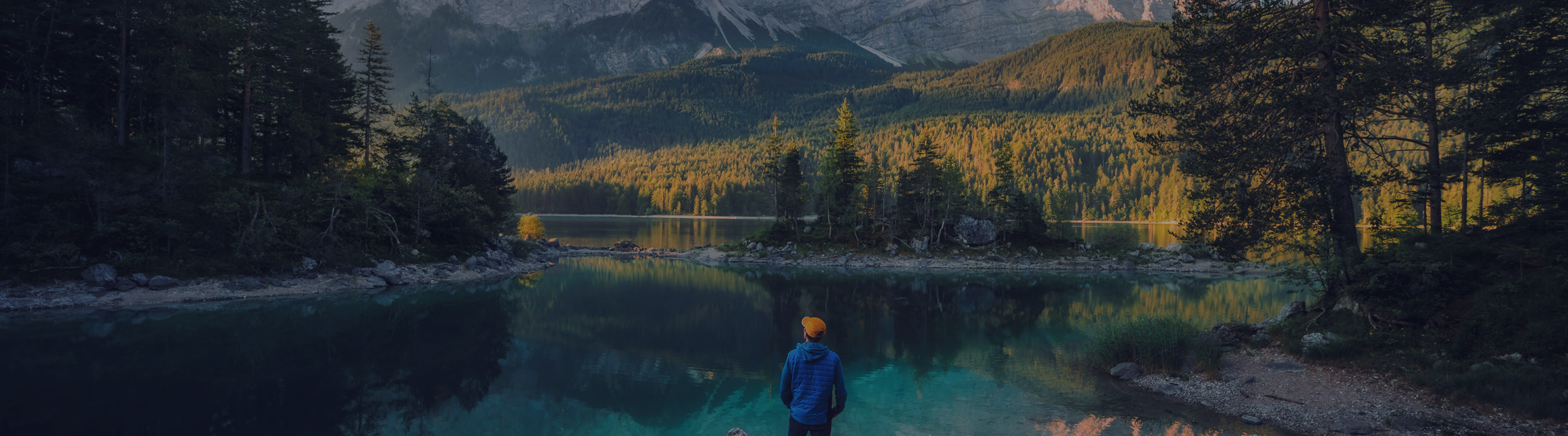 Person in blue jacket standing at the edge of a reflective alpine lake surrounded by forest during sunrise
