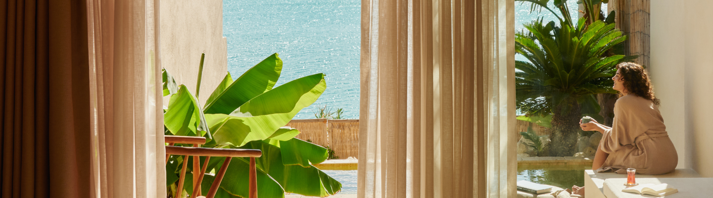 Hotel room with open doors to a terrace overlooking the sea, where a guest sits and relaxes in the sun.