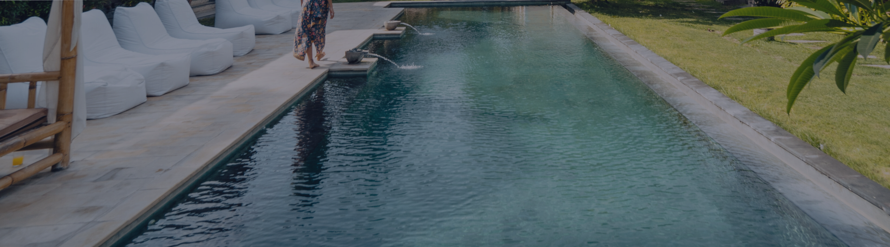 A person in a floral dress steps onto the edge of a modern outdoor swimming pool with white lounge chairs and manicured lawn nearby