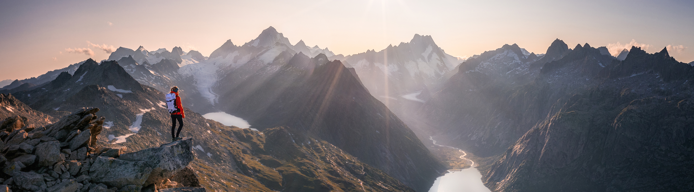 Solo hiker standing on a rocky ridge overlooking a dramatic mountain range and valley at sunset.