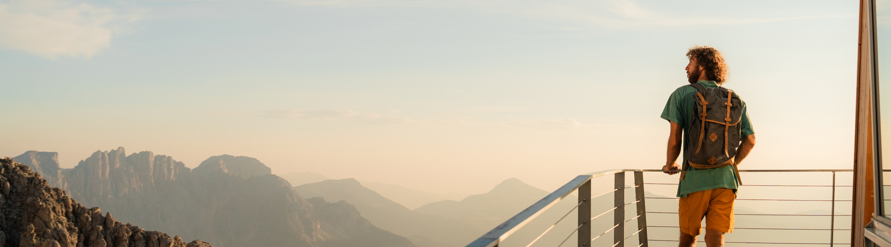 Traveler with a backpack standing on a viewing platform, looking out over a hazy mountain range at sunset.