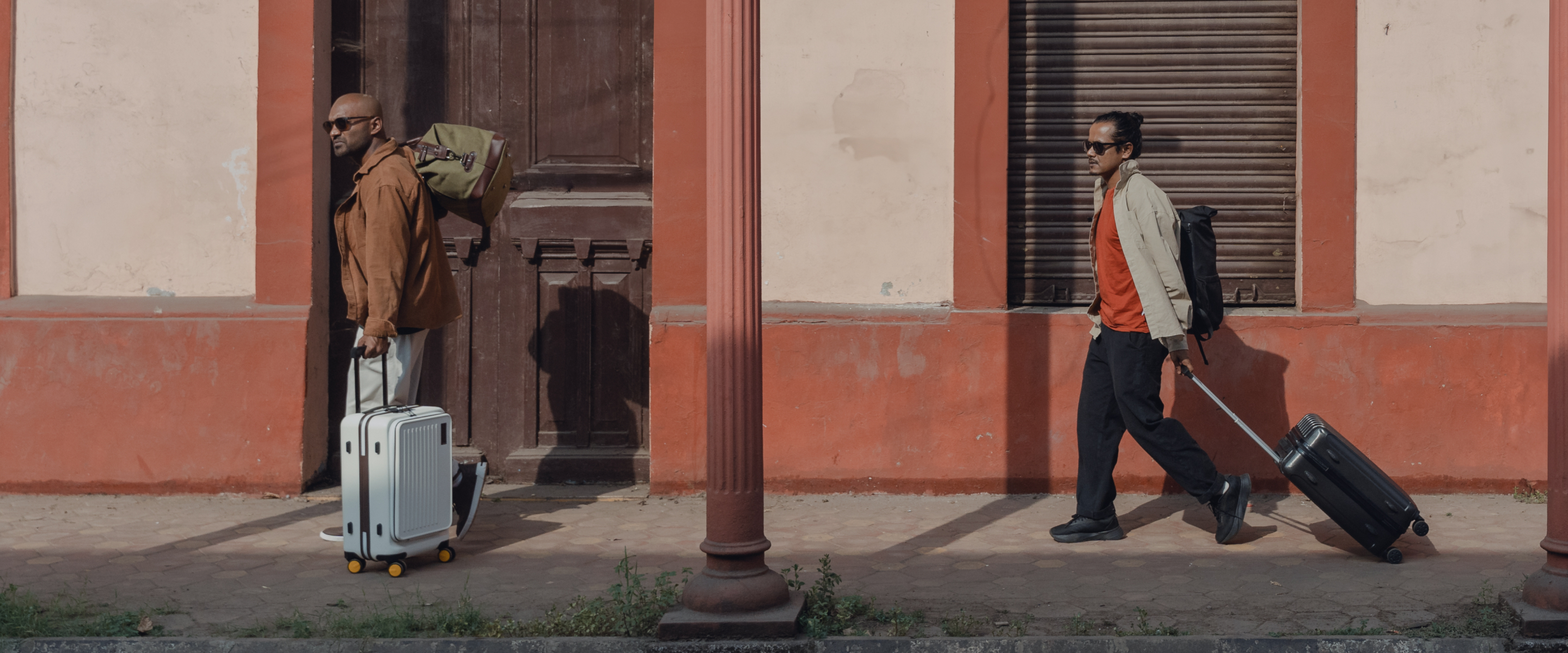 Two men are each pulling rolling suitcases while walking side by side along a sidewalk in front of a worn, reddish building with tall pillars. One man wears sunglasses and carries a duffel bag on his shoulder, while the other wears a light jacket and walks with a purposeful stride.