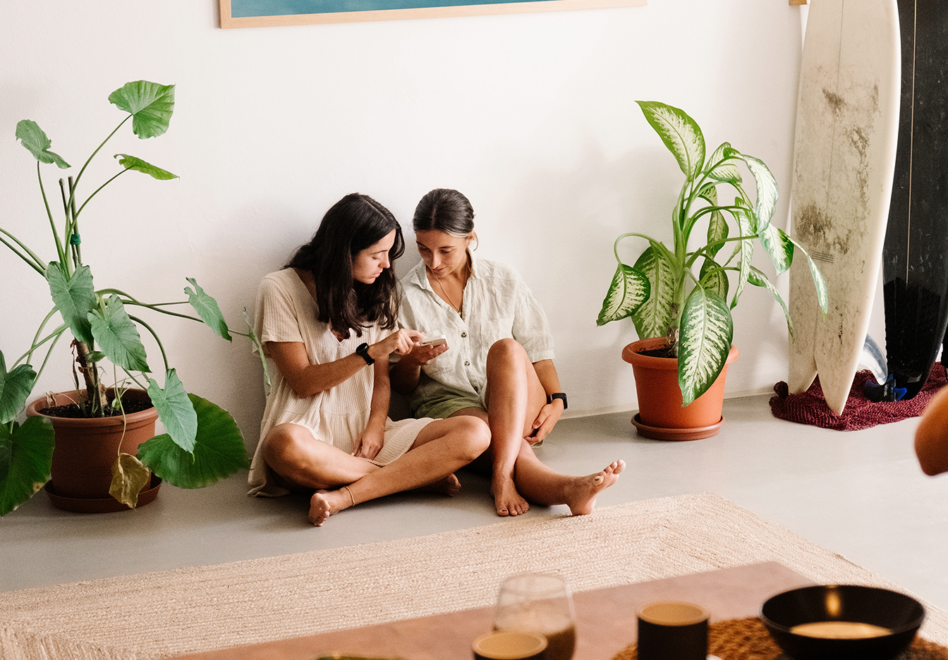 Two women sitting on the floor of a cozy room with potted plants and a surfboard, looking at a phone together.