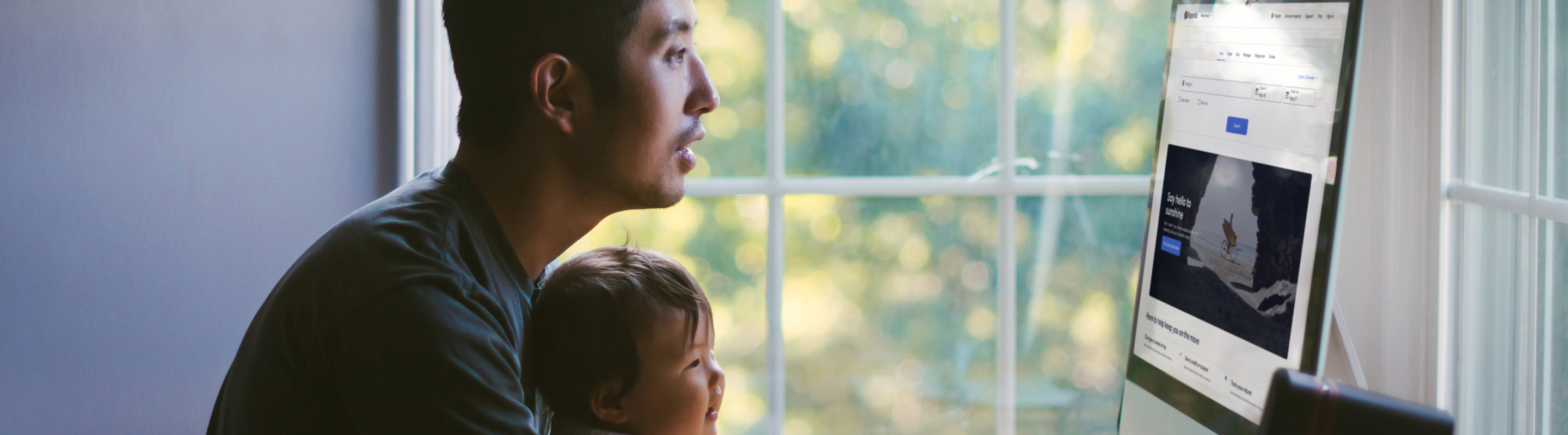 Dad booking travel online at home with baby on his lap, smiling and looking at computer screen.
