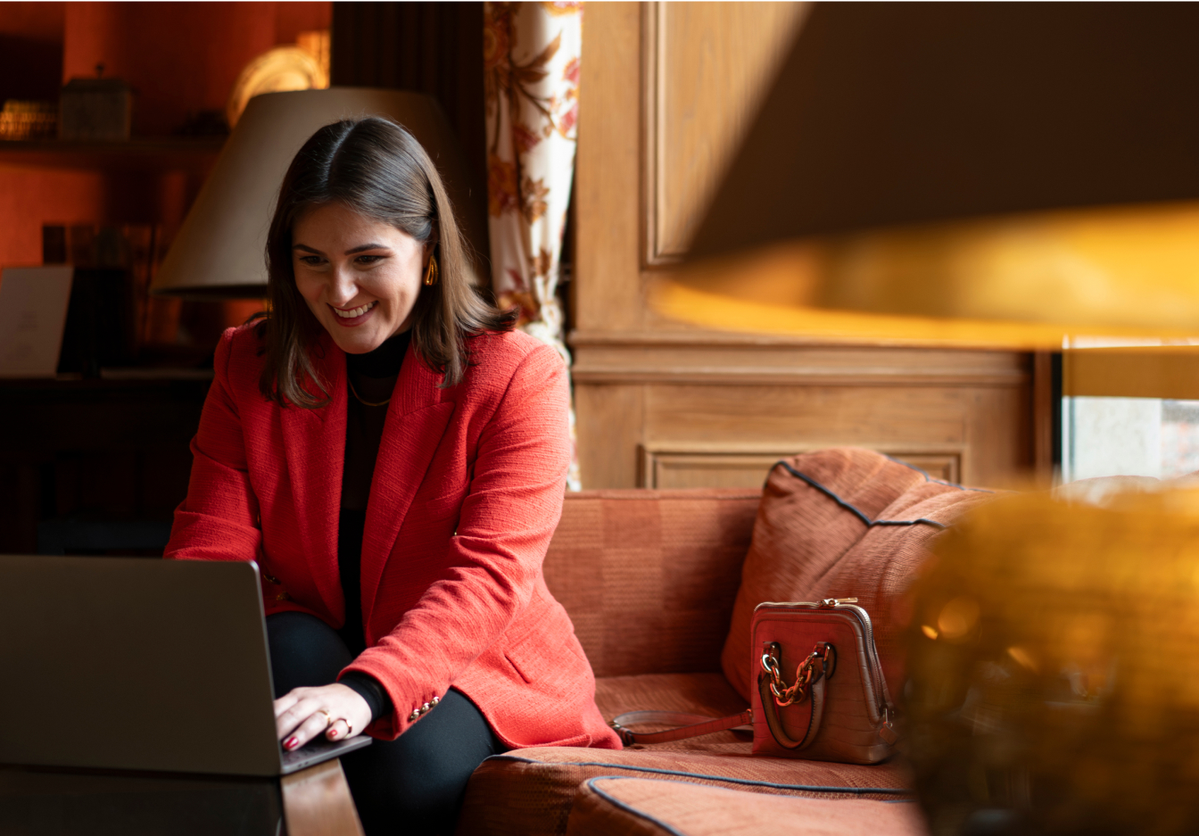 Woman in a red blazer smiling at her laptop while sitting in a warmly lit hotel room.