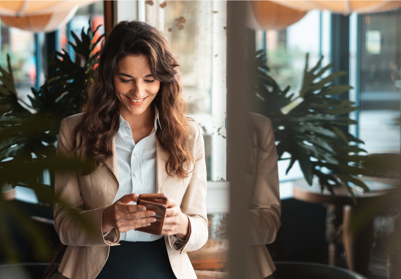Smiling woman looking down at her phone in a warmly lit space near a window, surrounded by indoor plants.