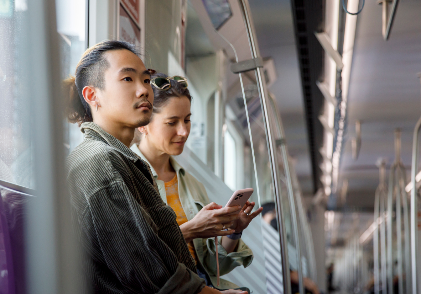 Two young adults seated on a subway train, one using a smartphone.